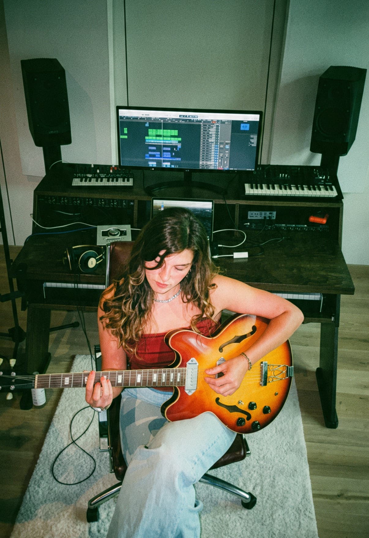 Vanessa Hartmann playing a sunburst semi-hollow guitar at her studio desk, with monitors, synthesizers and a screen behind her
